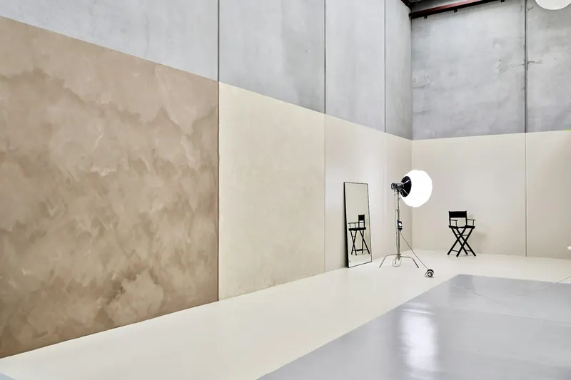 Le grand studio de l’atelier : murs de béton clair, un projecteur à boule, un miroir et une chaise de plateau