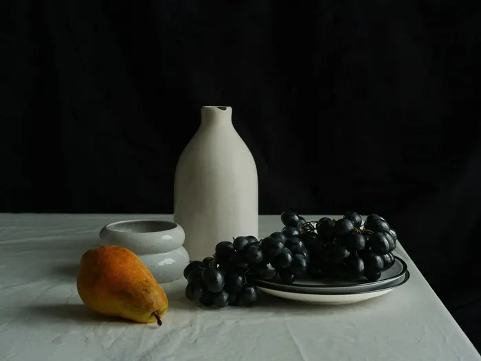 Nature morte : un vase blanc, une poire et une grappe de raisin noir sur une nappe, devant un fond sombre