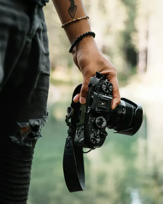 Une main tient un appareil photo hybride par sa sangle, au bord d’un lac
